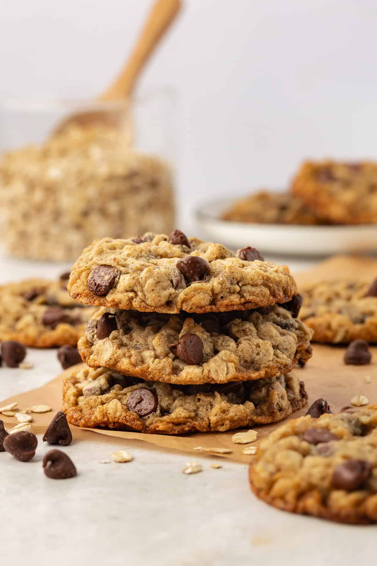a stack of three oatmeal chocolate chip cookies on brown parchment paper surrounded by more cookies, oats and chocolate chips scattered around