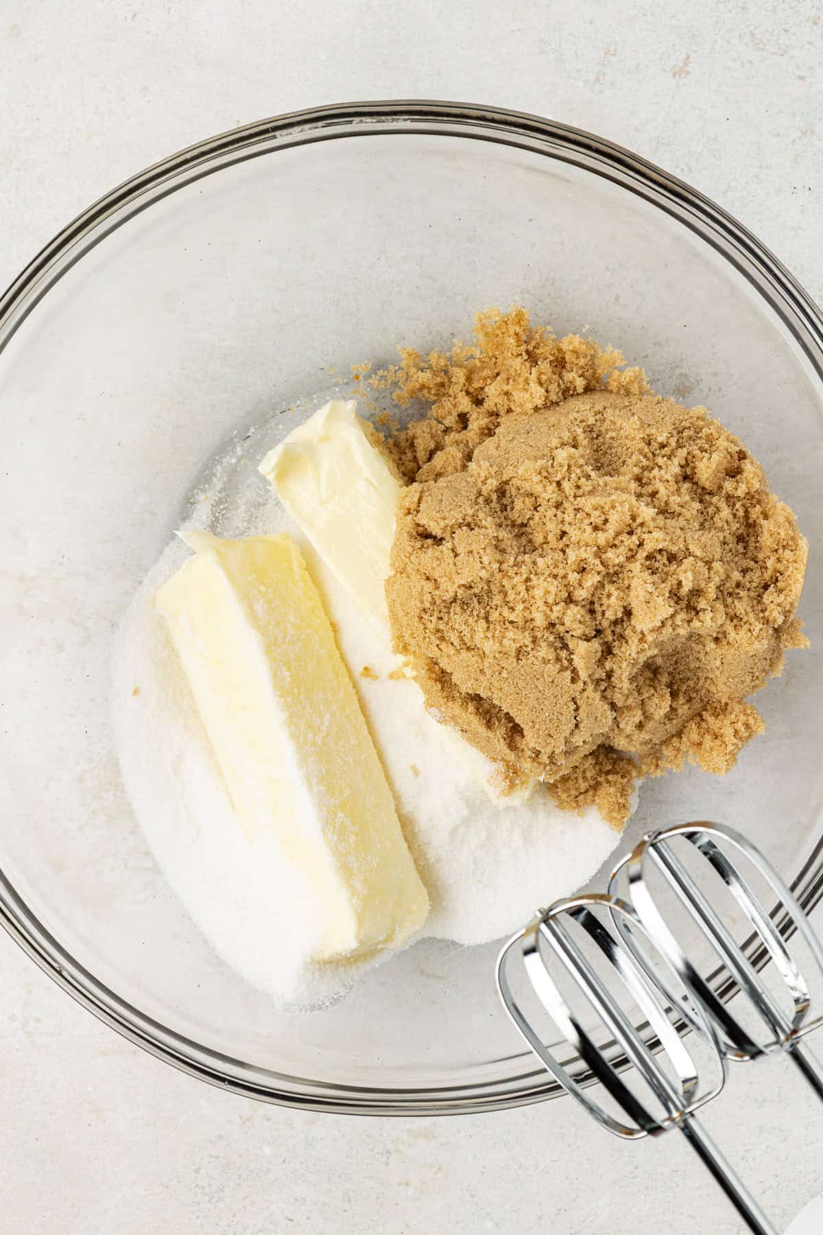 butter, granulated sugar and brown sugar in a clear glass mixing bowl with an electric mixer leaning on the side of the bowl