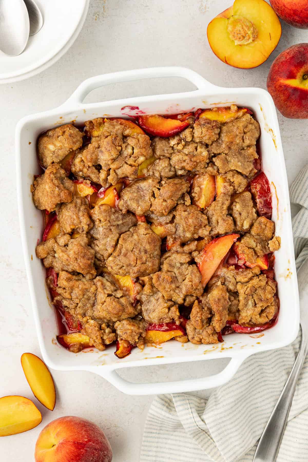 over head view of peach cobbler in a square white baking dish surrounded by fresh peaches, whole and slices, a stack of plates and spoon and a white and grey striped kitchen towel