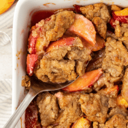 close up of peach cobbler in a white baking dish with a spoon stuck in it and fresh peaches to the left of the dish