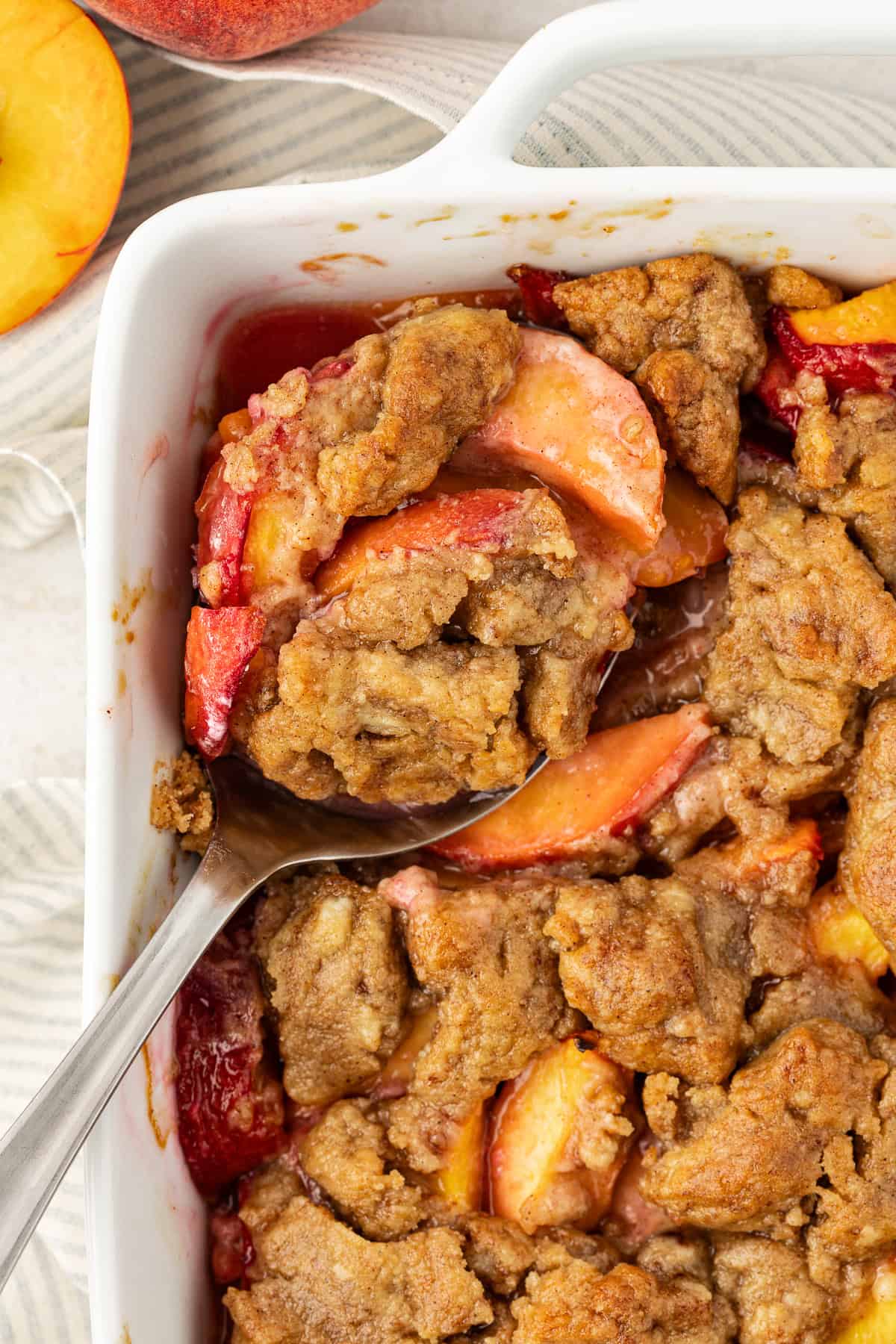 close up of peach cobbler in a white baking dish with a spoon stuck in it and fresh peaches to the left of the dish