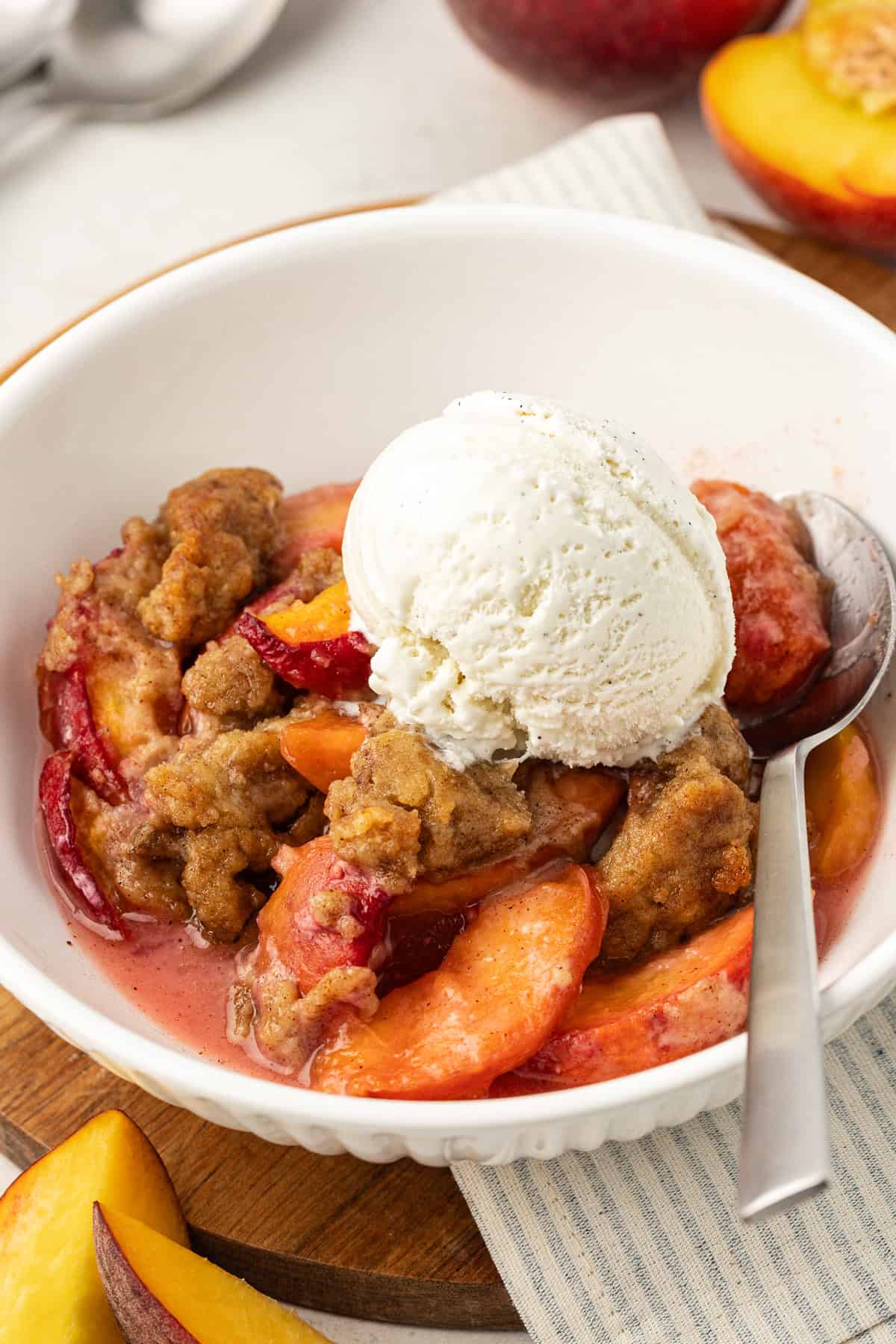 a serving of peach cobbler in a white bowl with a scoop of vanilla ice cream and a spoon, sitting on top of a wood platter with a white and grey striped kitchen towel and surrounded by fresh peach slices