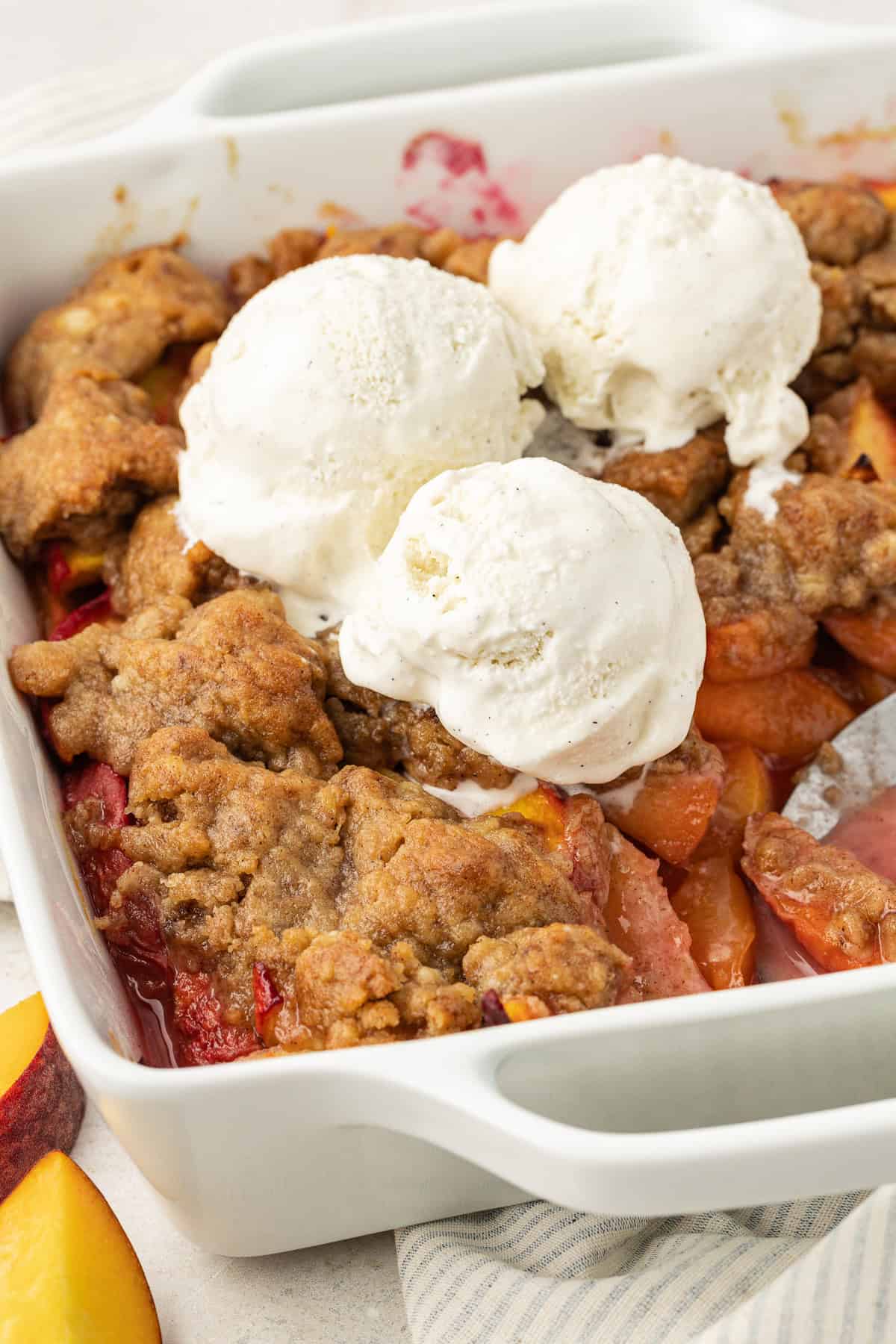 peach cobbler in a square white baking dish topped with three scoops of vanilla ice cream