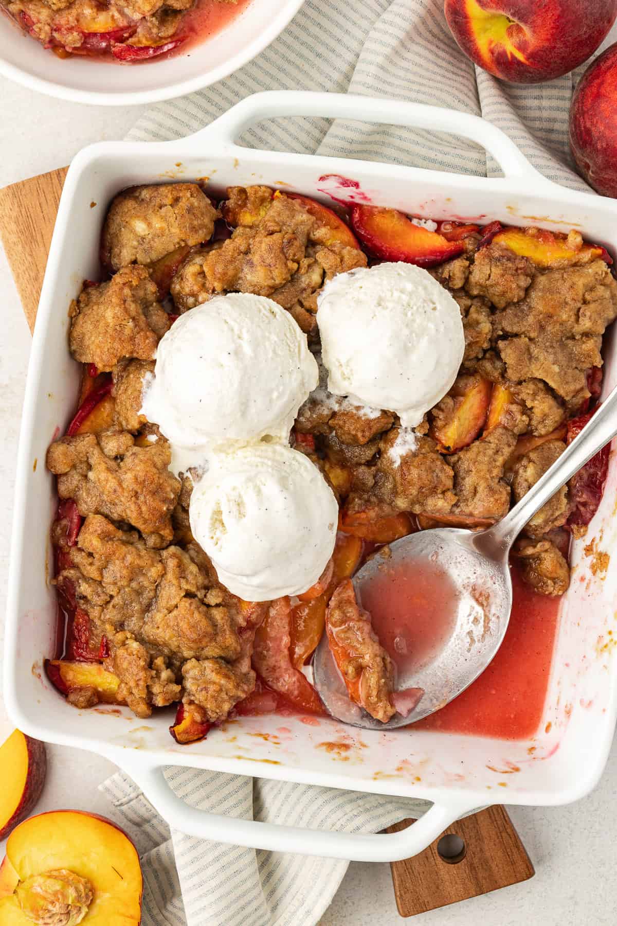 over head view of a baking dish of peach cobbler topped with three scoops of vanilla ice cream, a scoop of cobbler missing with a spoon laying in the dish, surrounded by fresh peaches