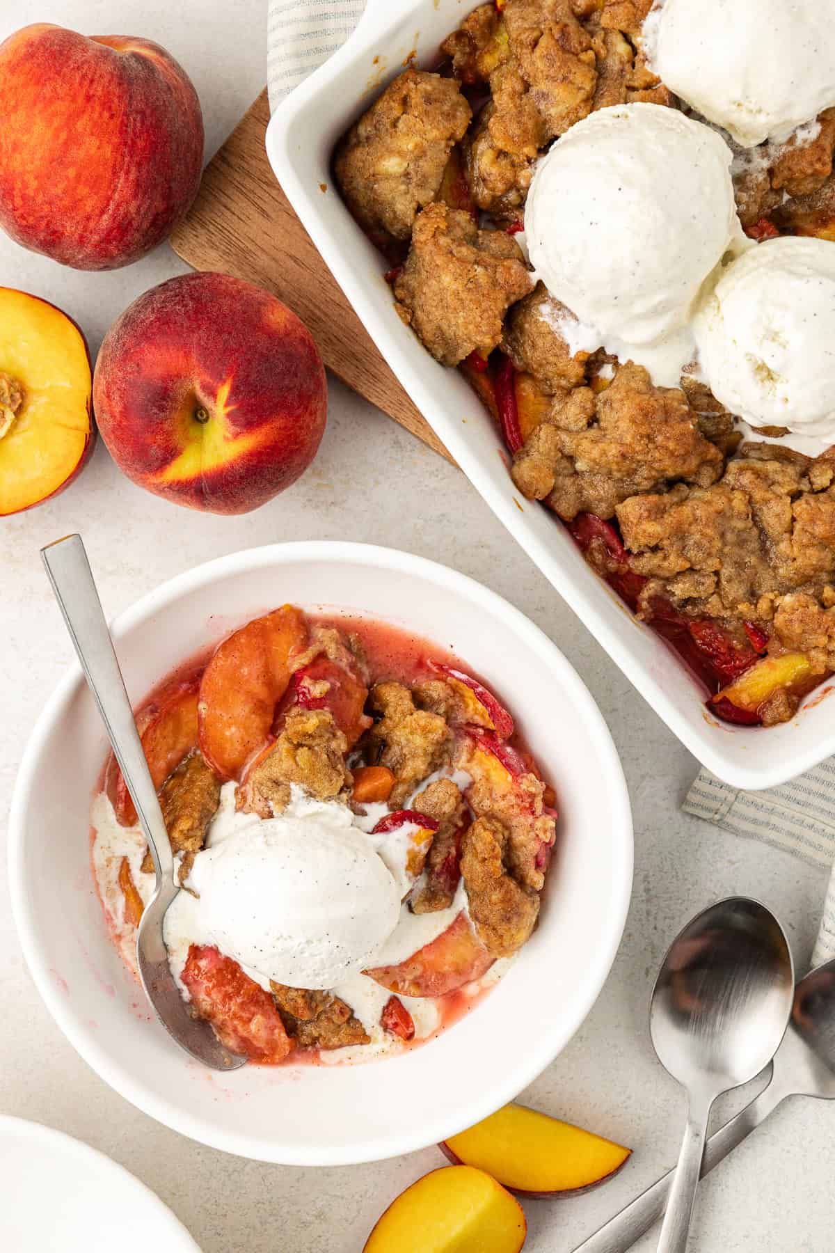 over head view of a bowl of peach cobbler with a scoop of vanilla ice cream and a spoon surrounded by the baking dish of cobbler with ice cream on top, fresh peaches whole and sliced, and two spoons