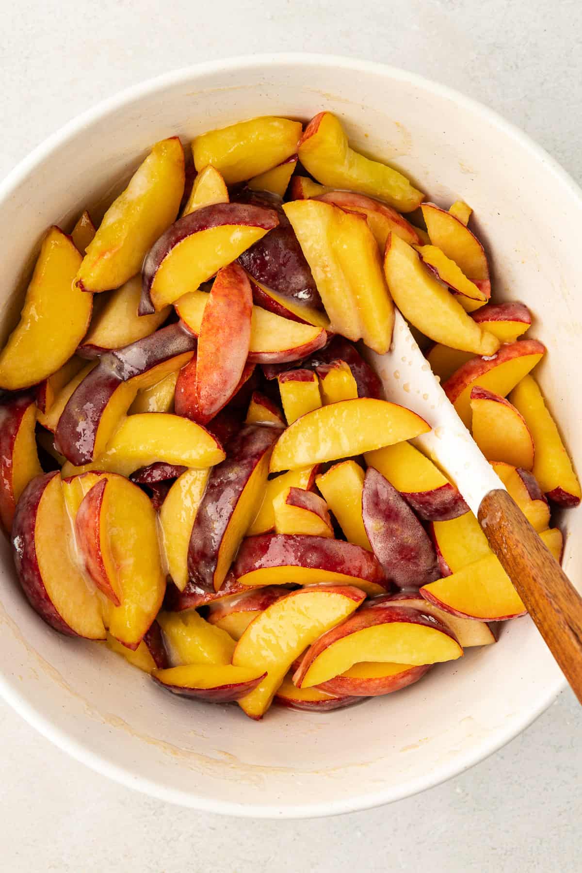 white bowl full of fresh peach slices being mixed with ingredients for peach cobbler with a white spatula