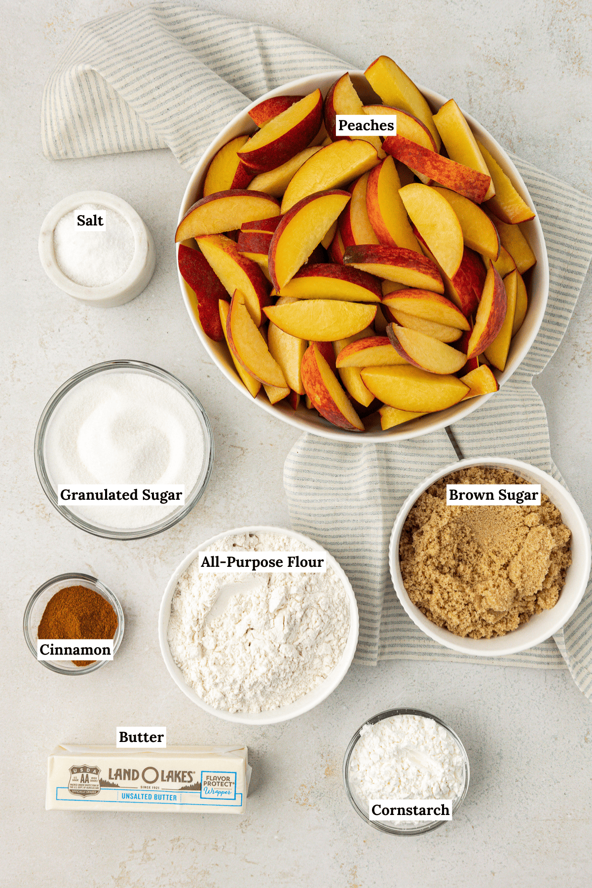 over head view of the ingredients for peach cobbler including sliced peaches, salt, granulated sugar, brown sugar, all-purpose flour, cinnamon, butter and cornstarch