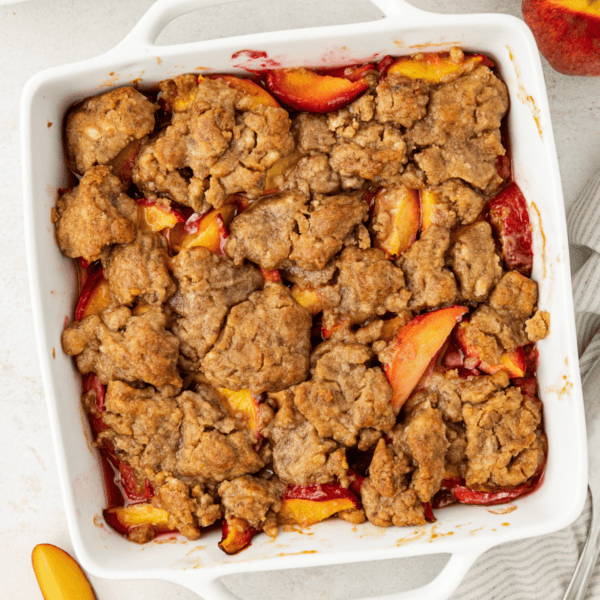 over head view of peach cobbler in a square white baking dish surrounded by fresh peaches, whole and slices, a stack of plates and spoon and a white and grey striped kitchen towel