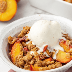 a small white bowl of peach crisp with a scoop of vanilla ice cream on top of a light purple kitchen towel with fresh peaches and the baking dish of peach crisp in the background