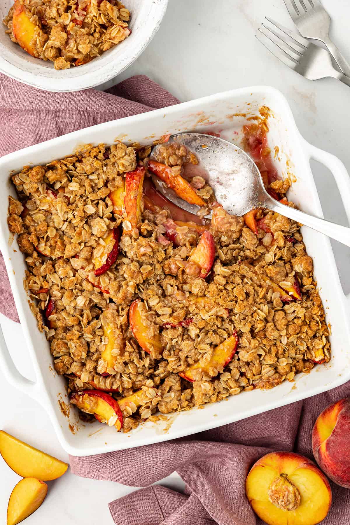 over head view of a baking dish of peach crisp with a spoon in it and a scoop missing, sitting on top of a light purple towel and surrounded by a bowl of peach crisp, forks, and fresh peaches sliced and whole