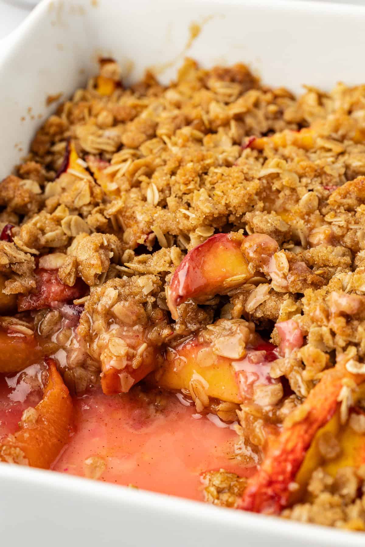 close up of peach crisp in a white baking dish with a scoop missing