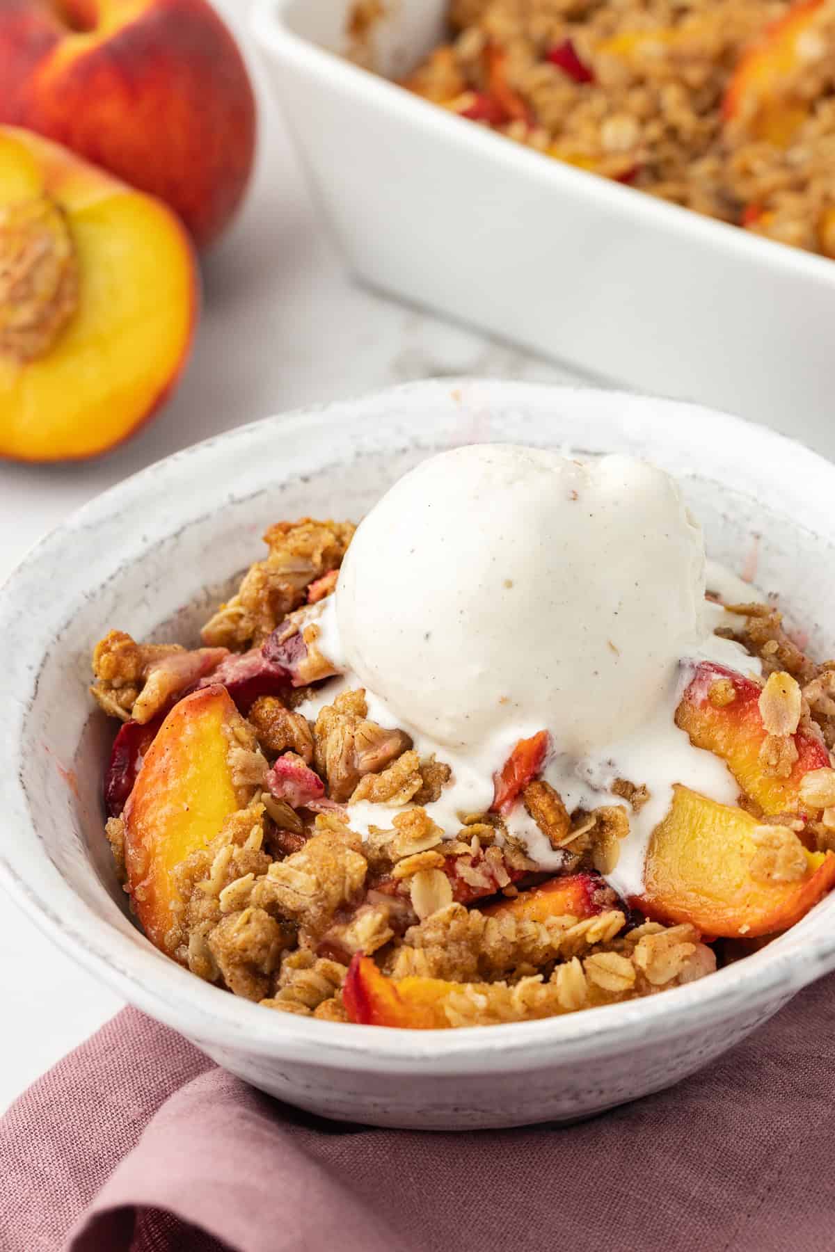 a small white bowl of peach crisp with a scoop of vanilla ice cream on top of a light purple kitchen towel with fresh peaches and the baking dish of peach crisp in the background