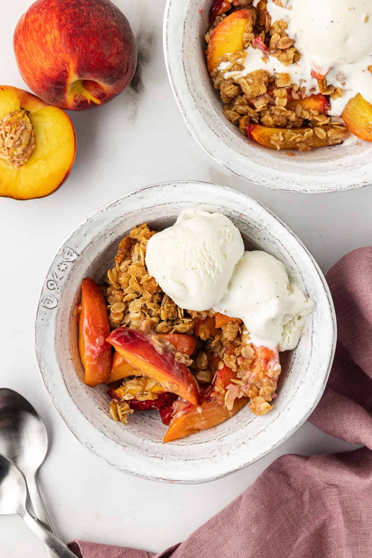 two white bowls of peach crisp with scoops of vanilla ice cream, a light purple kitchen towels, two spoons and fresh peaches