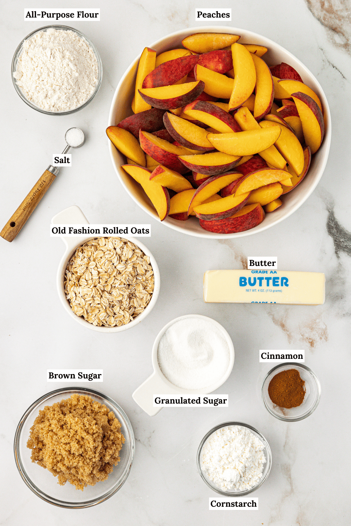 over head view of peach crisp ingredients including all-purpose flour, fresh sliced peaches, salt, old fashioned rolled oats, butter, brown sugar, granulated sugar, cinnamon, and cornstarch