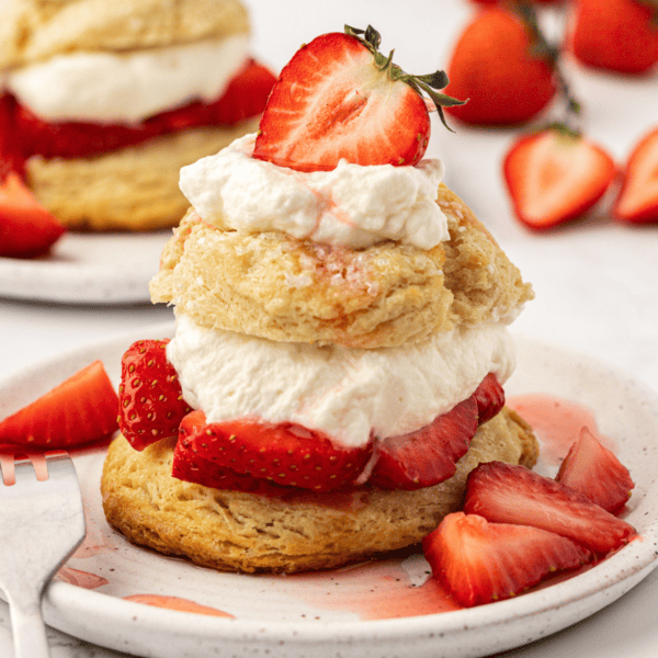 strawberry shortcake on a small white plate with a fork and more strawberries sprinkled around and another plate of strawberry shortcake in the background