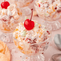 a small trifle dish full of ambrosia salad with a cherry on top surrounded by more dishes of salad, a light orange towel and a spoon