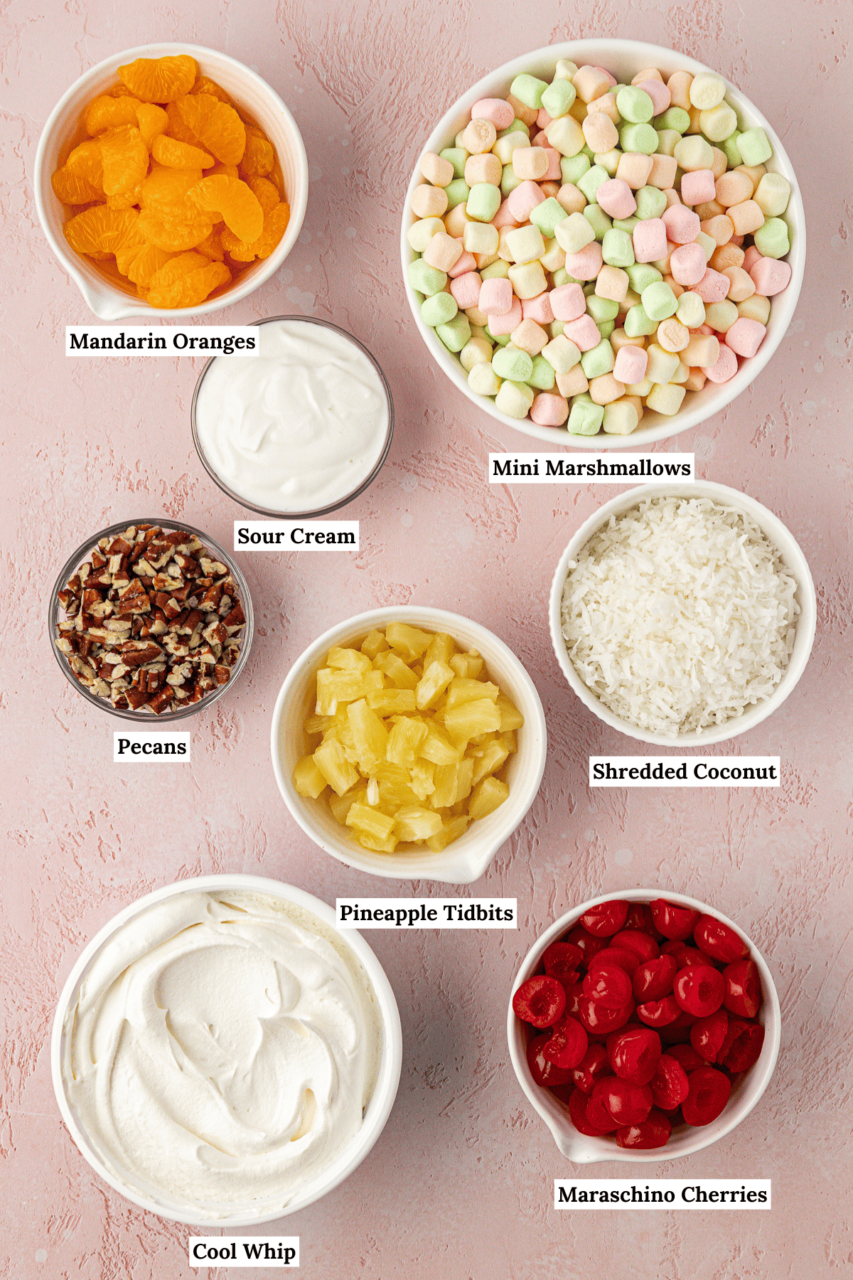 over head view of the ingredients for ambrosia salad including mandarin oranges, sour cream, mini marshmallows, pecans, pineapple tidbits, shredded coconut, cool whip and maraschino cherries