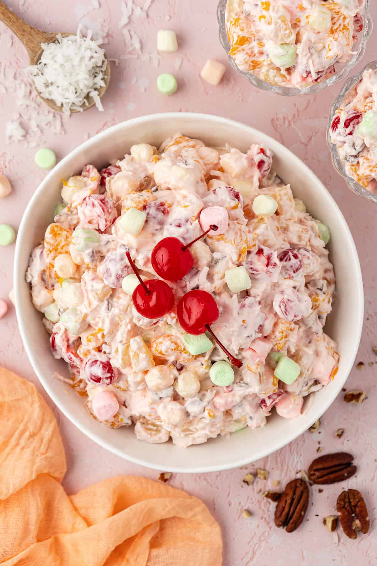 over head view of a large white bowl of ambrosia salad topped with 3 cherries, surrounded by two small dishes of ambrosia, a spoon of coconut flakes, a light orange towel and mini marshmallows and pecans scattered around