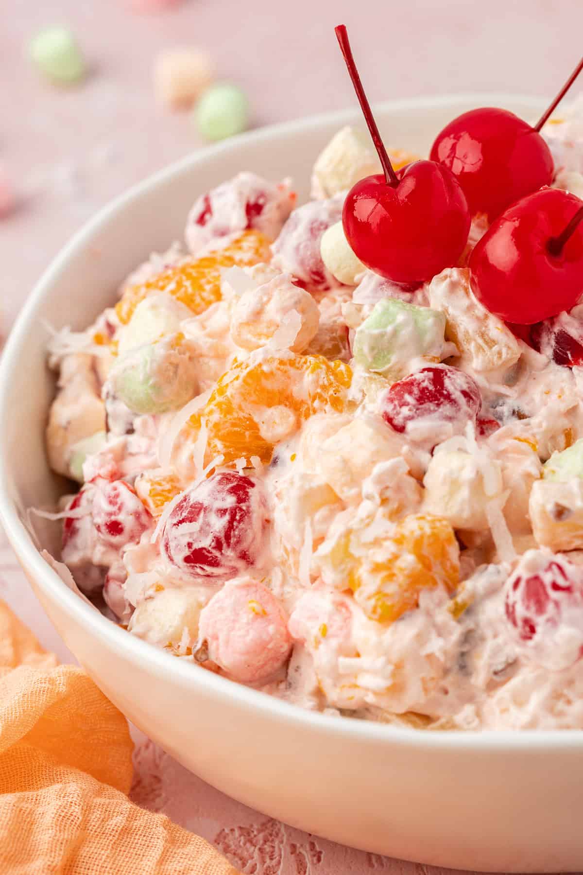 close up of a white bowl full of ambrosia salad topped with 3 cherries, with a light orange towel beside the bowl and mini marshmallows scattered around