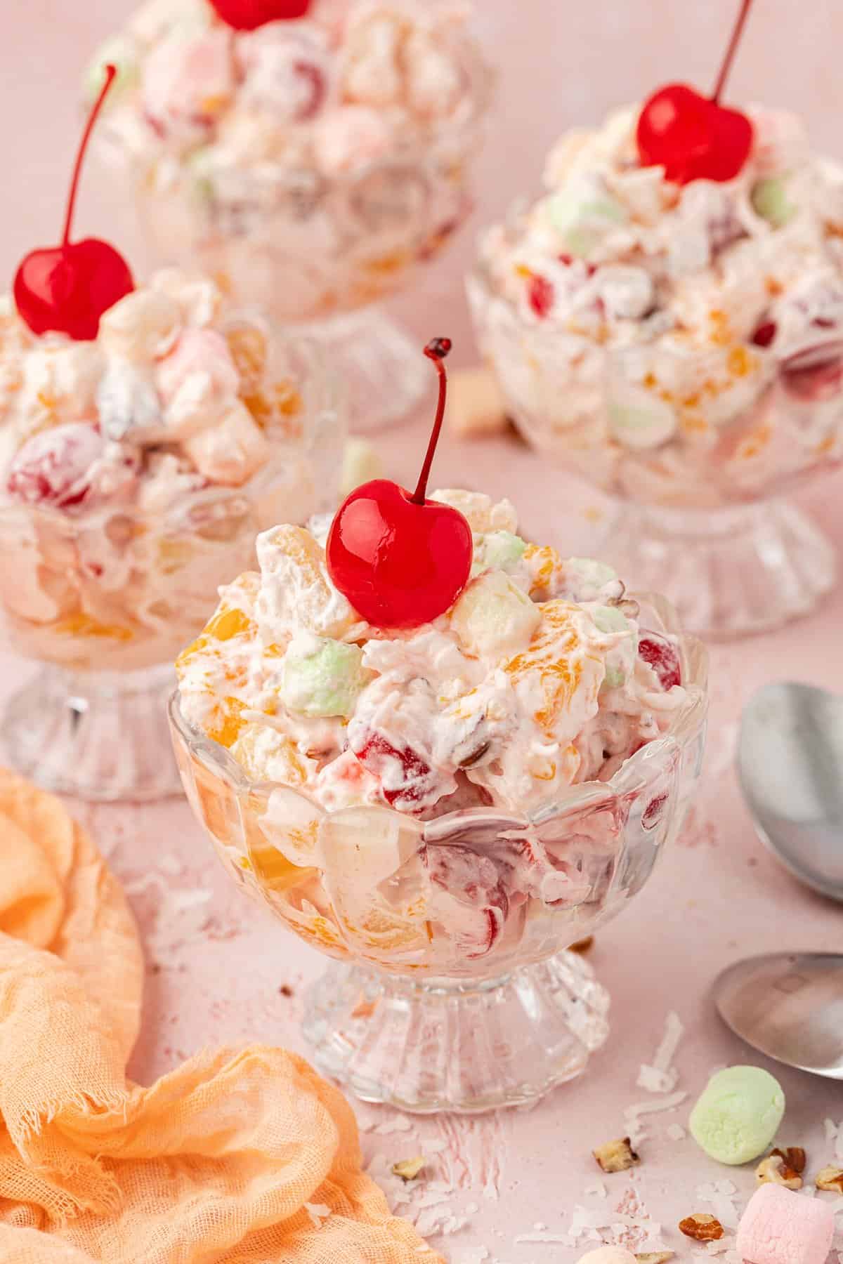 4 small trifle dishes of ambrosia salad topped with cherries with a light orange towel and spoons beside them and mini marshmallows, shredded coconut and little pecan bits scattered around