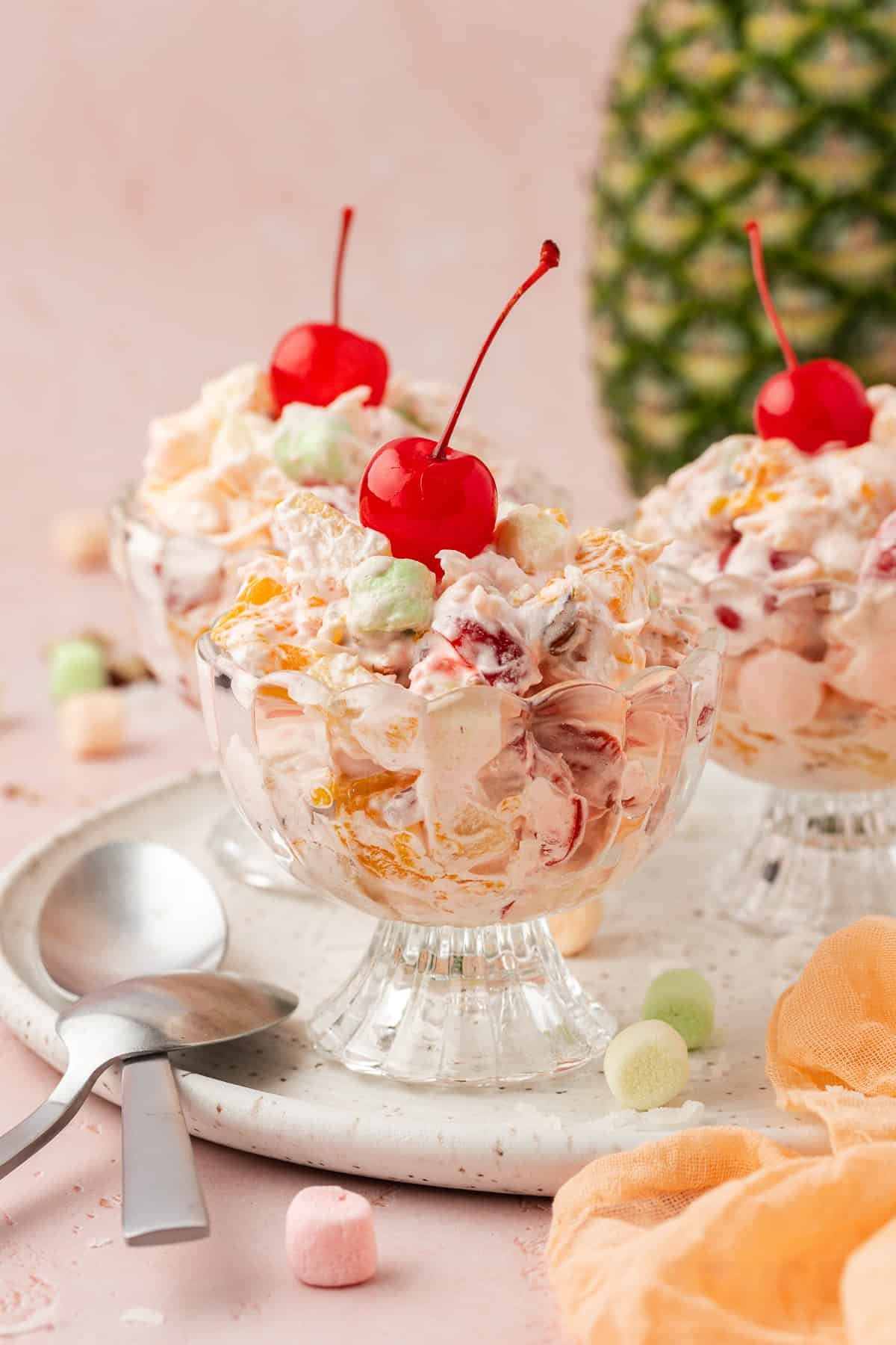 three small trifle dishes of ambrosia salad topped with cherries on a white platter with spoons resting on it, a whole pineapple in the background, a light orange towel and mini marshmallows scattered around