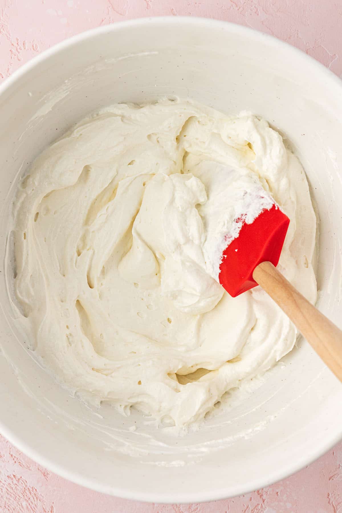 sour cream and whipped topping being mixed with a spatula in a white bowl on a pink surface