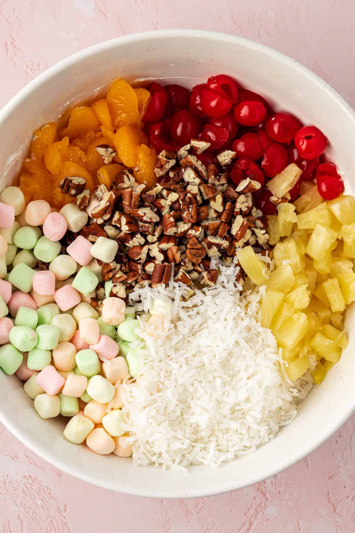 a large white bowl with the ingredients for ambrosia salad that have not been mixed together yet including mini marshmallows, mandarin oranges, maraschino cherries, chopped pecans, pineapple tidbits and coconut flakes