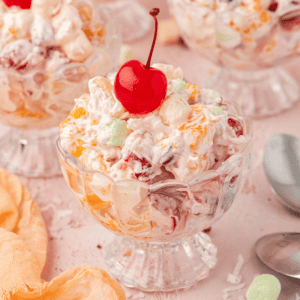 a small trifle dish full of ambrosia salad with a cherry on top surrounded by more dishes of salad, a light orange towel and a spoon