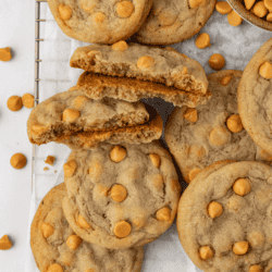 over head view of a pile of butterscotch cookies on a wire rack covered in white parchment paper with a bowl of butterscotch chips and more butterscotch chips scattered around