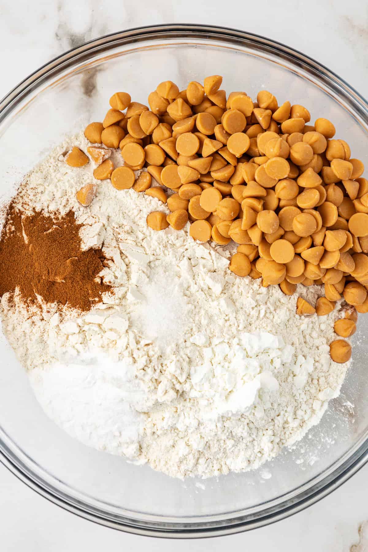 dry ingredients for butterscotch cookies in a clear glass bowl, not mixed together yet showing the flour with sugar, cinnamon, cornstarch, baking soda and butterscotch chips on top