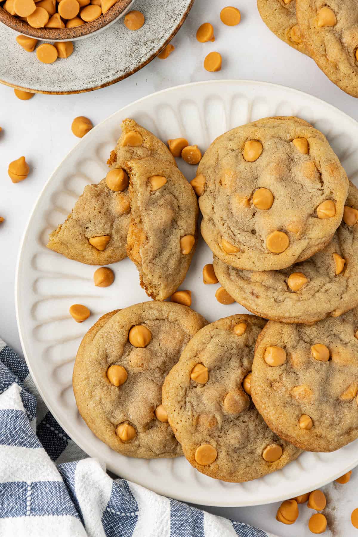 over head view of a plate piled with butterscotch cookies, most full but one broken in half, more cookies, a bowl of butterscotch chips and more butterscotch chips sprinkled around