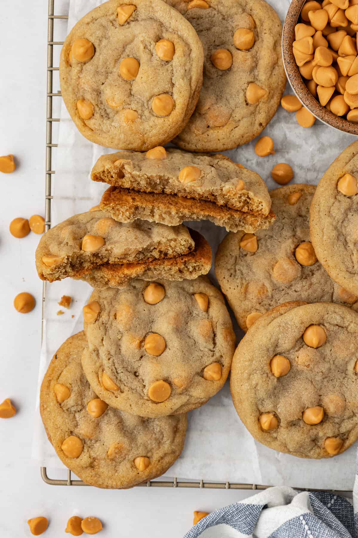 over head view of a pile of butterscotch cookies on a wire rack covered in white parchment paper with a bowl of butterscotch chips and more butterscotch chips scattered around