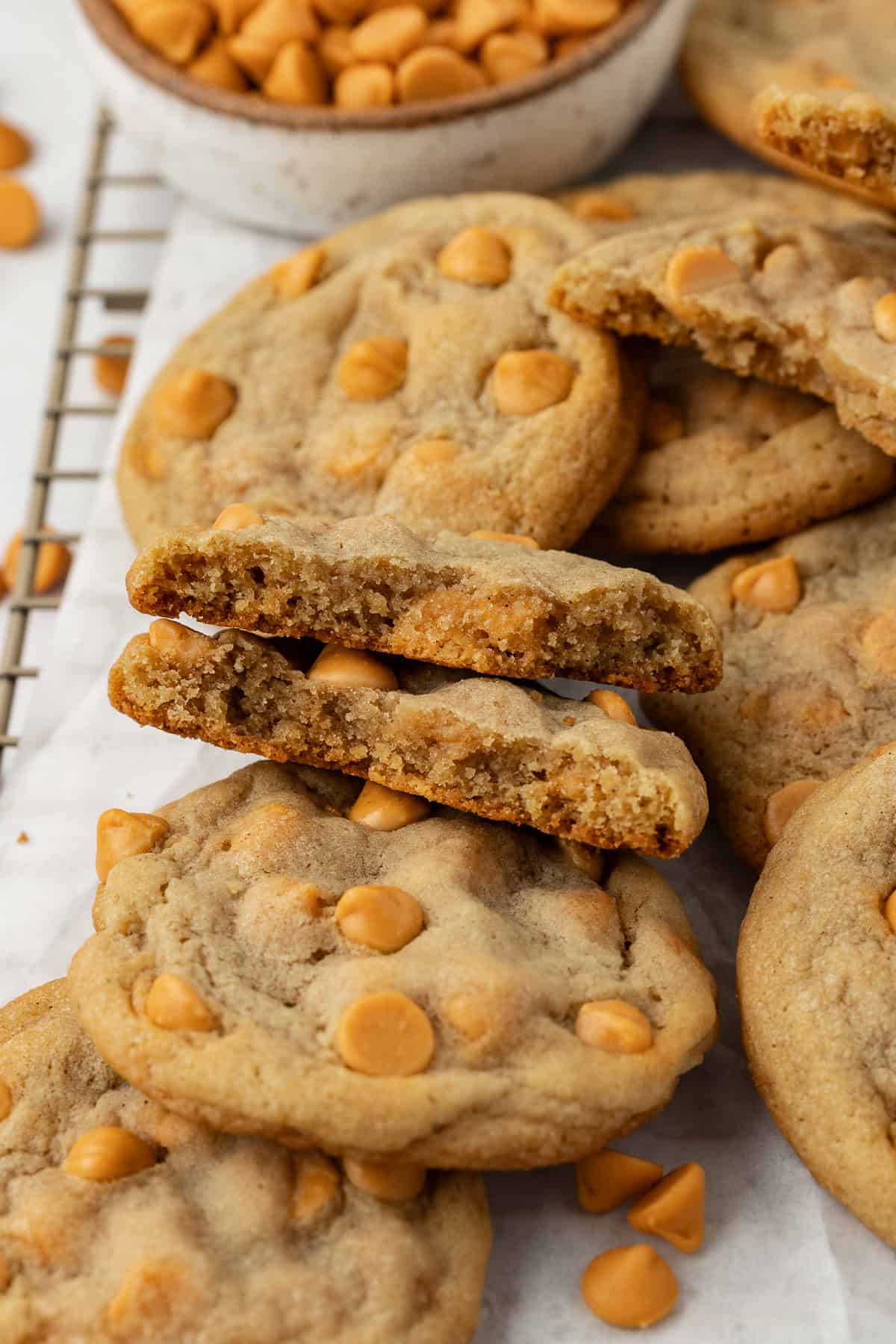 close up of a pile of butterscotch cookies on a wire rack lined with white parchment paper, with one cookie broken half showing the inside of the cookie and butterscotch chips sprinkled around