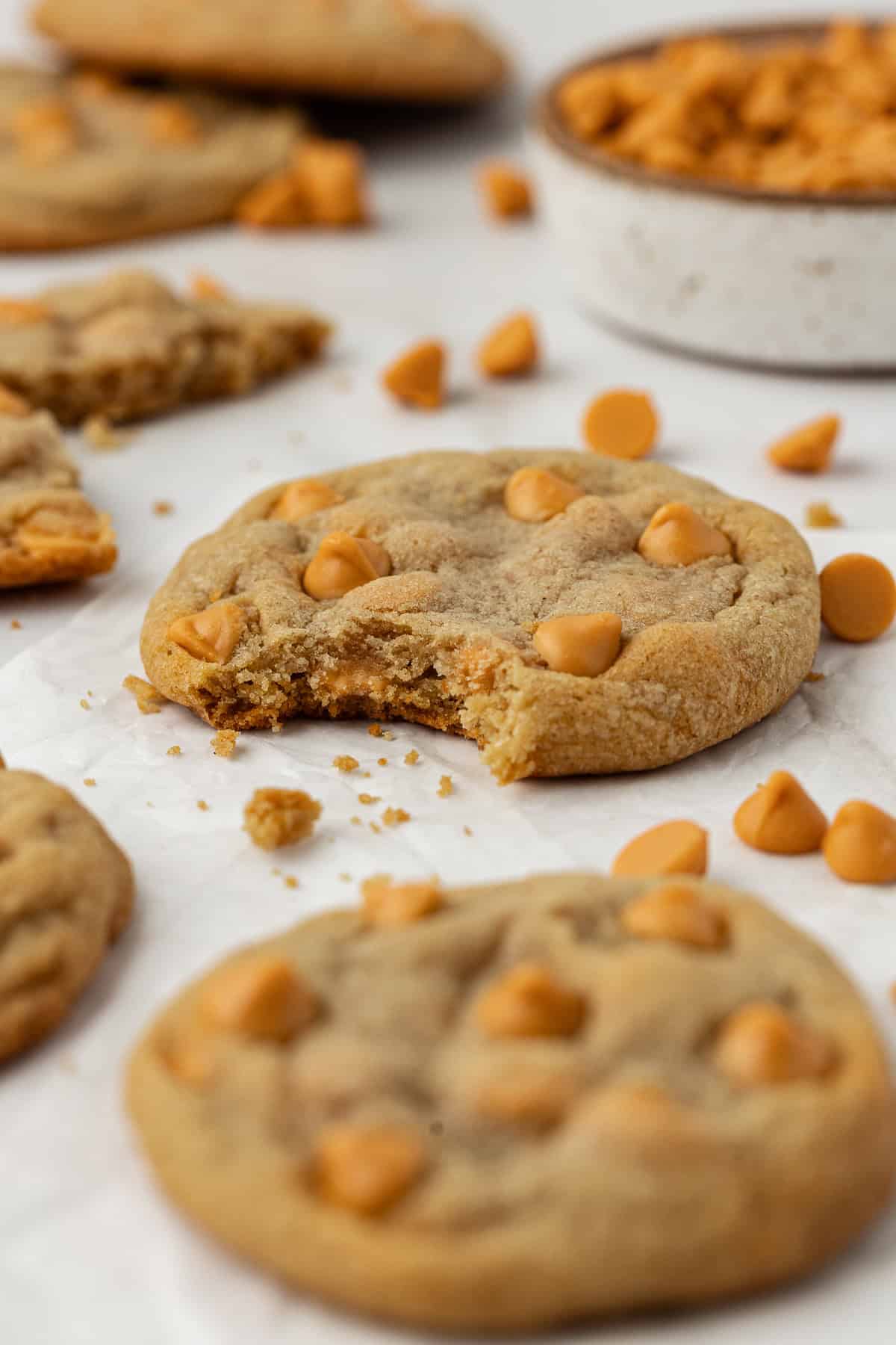 butterscotch cookies scattered on white parchment paper with a bite missing from the center cookie, cookie crumbs and butterscotch chips scattered around