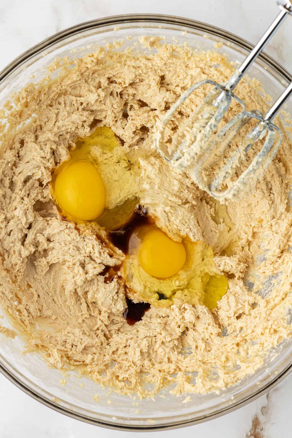 eggs and vanilla being combined with a creamed mixture of brown sugar and butter in a clear glass bowl with an electric mixer