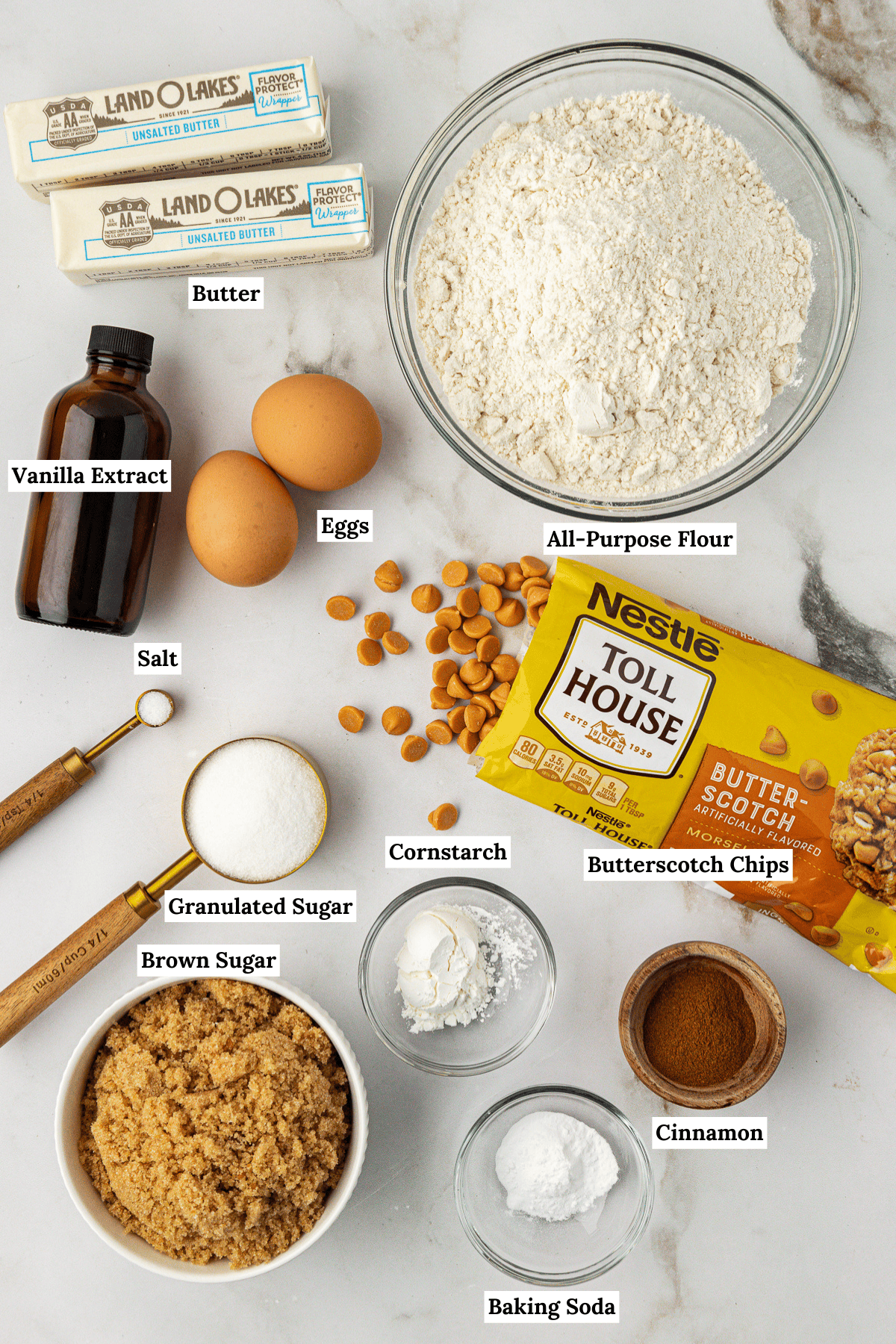 Over head view of the ingredients for butterscotch cookies including butter, vanilla extract, eggs, all-purpose flour, salt, granulated sugar, cornstarch, butterscotch chips, brown sugar, baking soda and cinnamon