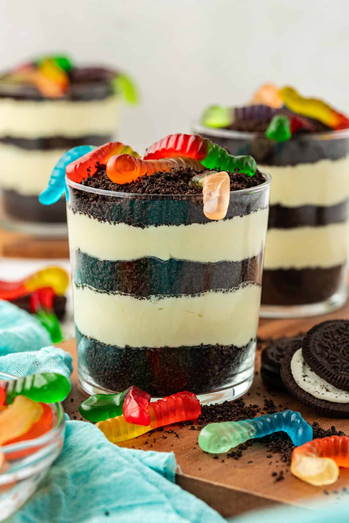 three small trifle dishes of dirt cake topped with gummy worms on a wood platter surrounded by oreos, crushed oreos, gummy worms and a light teal towel