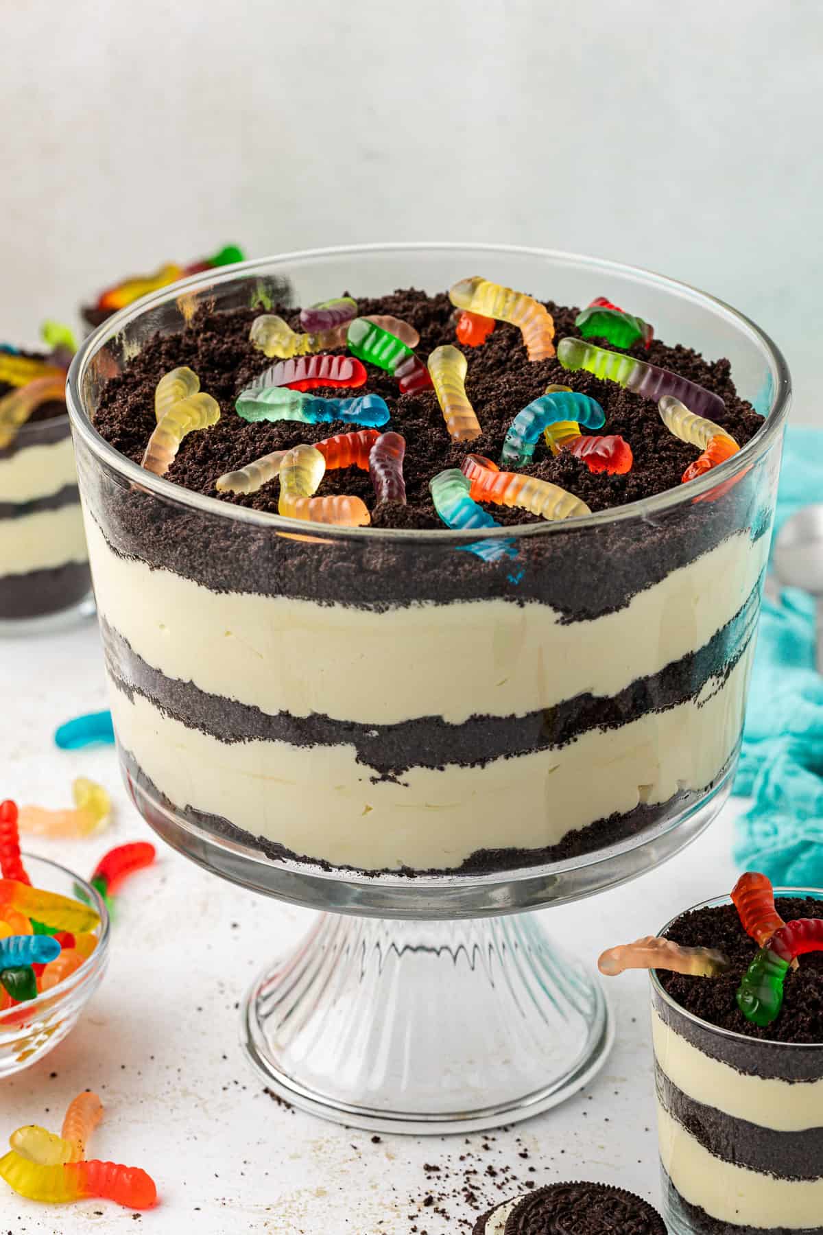 a large clear glass trifle dish full of dirt cake topped with gummy worms surrounded by smaller dirt cake trifle dishes, a teal towel, gummy worms and oreos