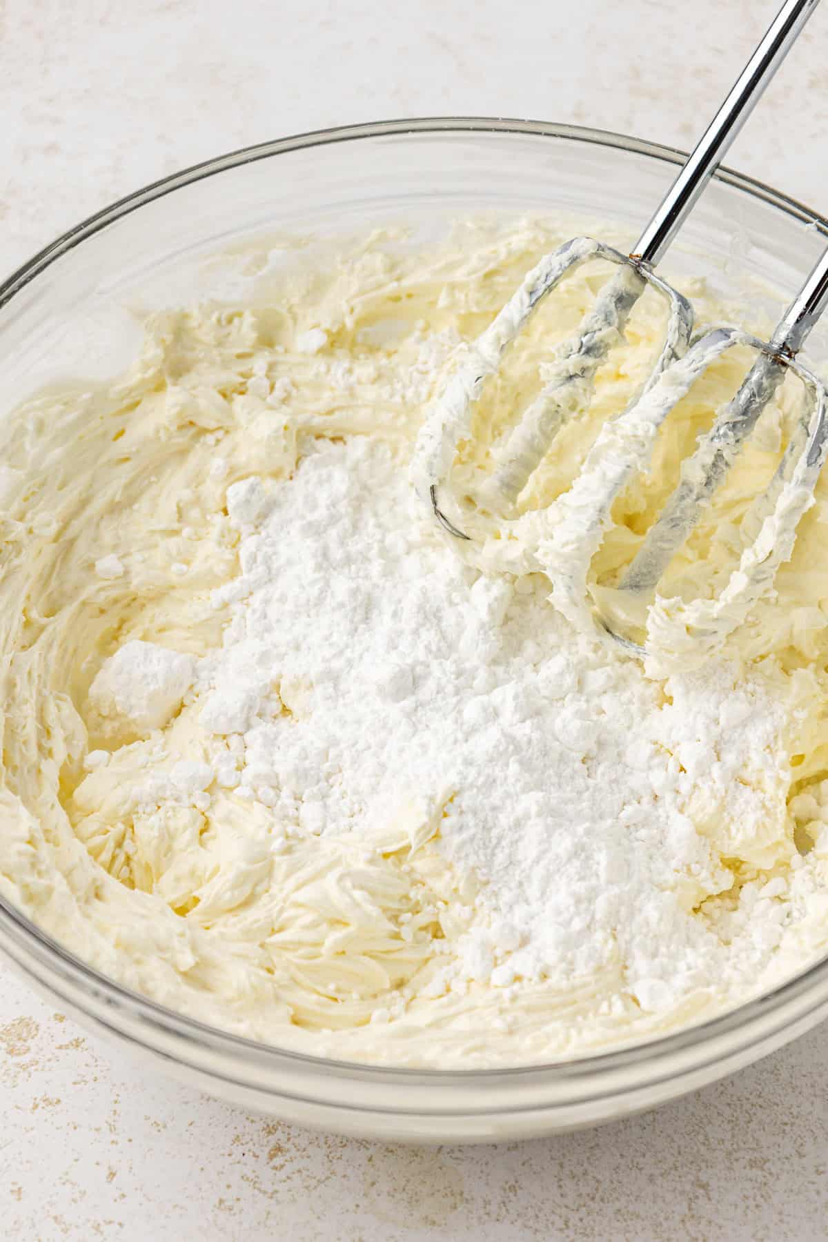 a cream cheese, butter and powdered sugar mixture being beaten in a large clear glass bowl with an electric mixer