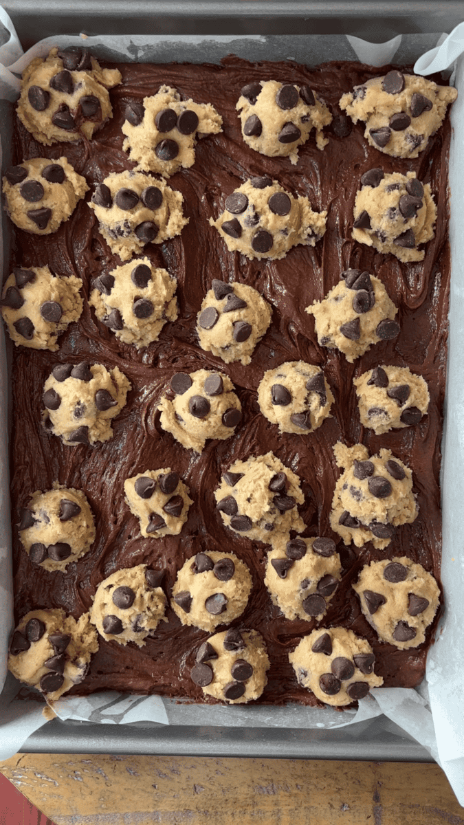 A baking pan filled with chocolate brownie batter, topped with multiple scoops of chocolate chip cookie dough, ready to be baked into delicious brookies.