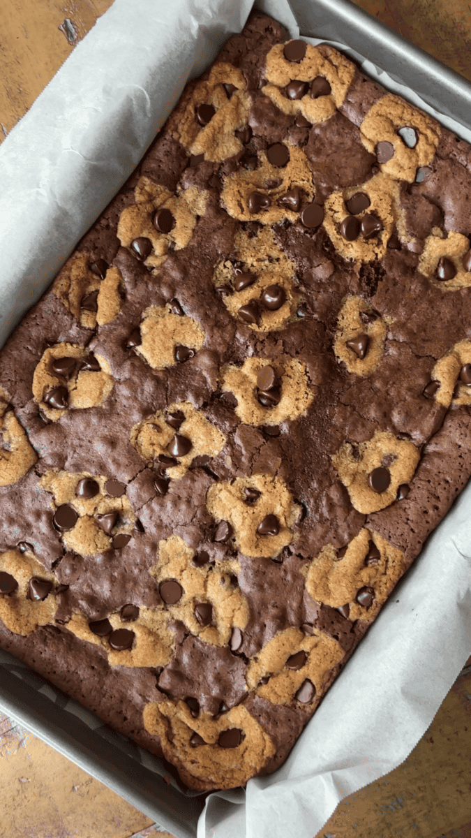 A baked brookie, the perfect fusion of brownie and chocolate chip cookie dough, is nestled in a parchment-lined pan. Chocolate chips are generously scattered on top, making these brookies an irresistible dessert.