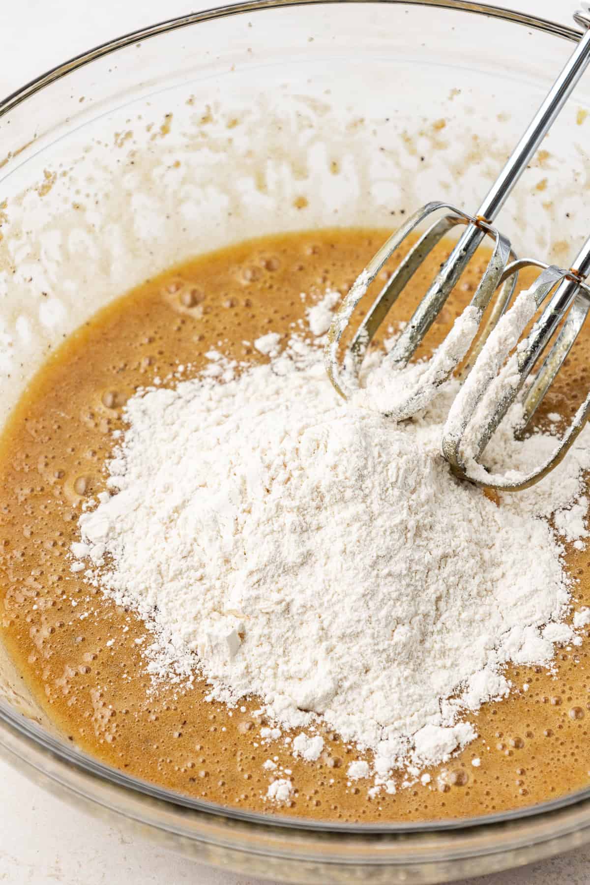 dry ingredients on top of wet ingredients for blondies in a clear glass bowl with an electric mixer leaning on the side