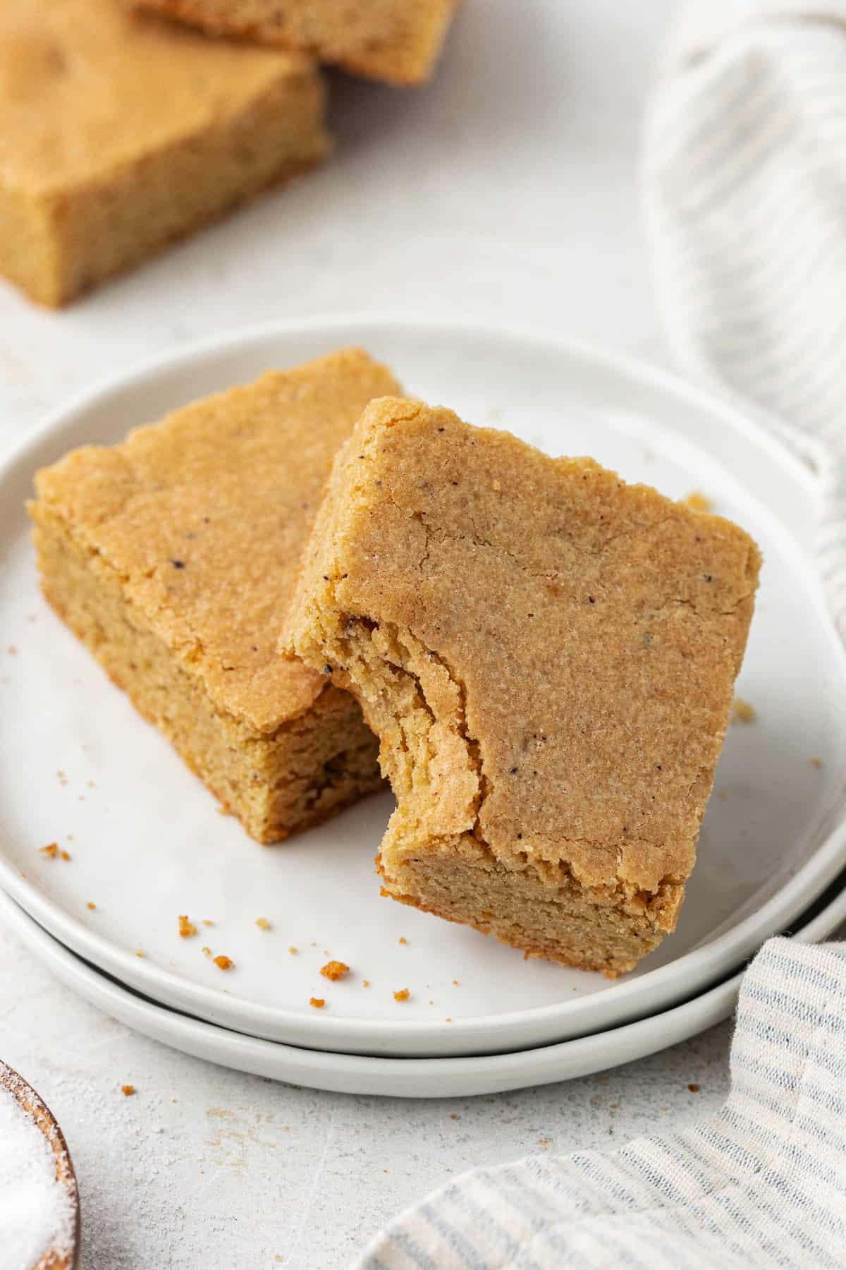 two blondie squares on a stack of two small white plates, with the top blondie missing a bite out of it and leaning on the other blondie