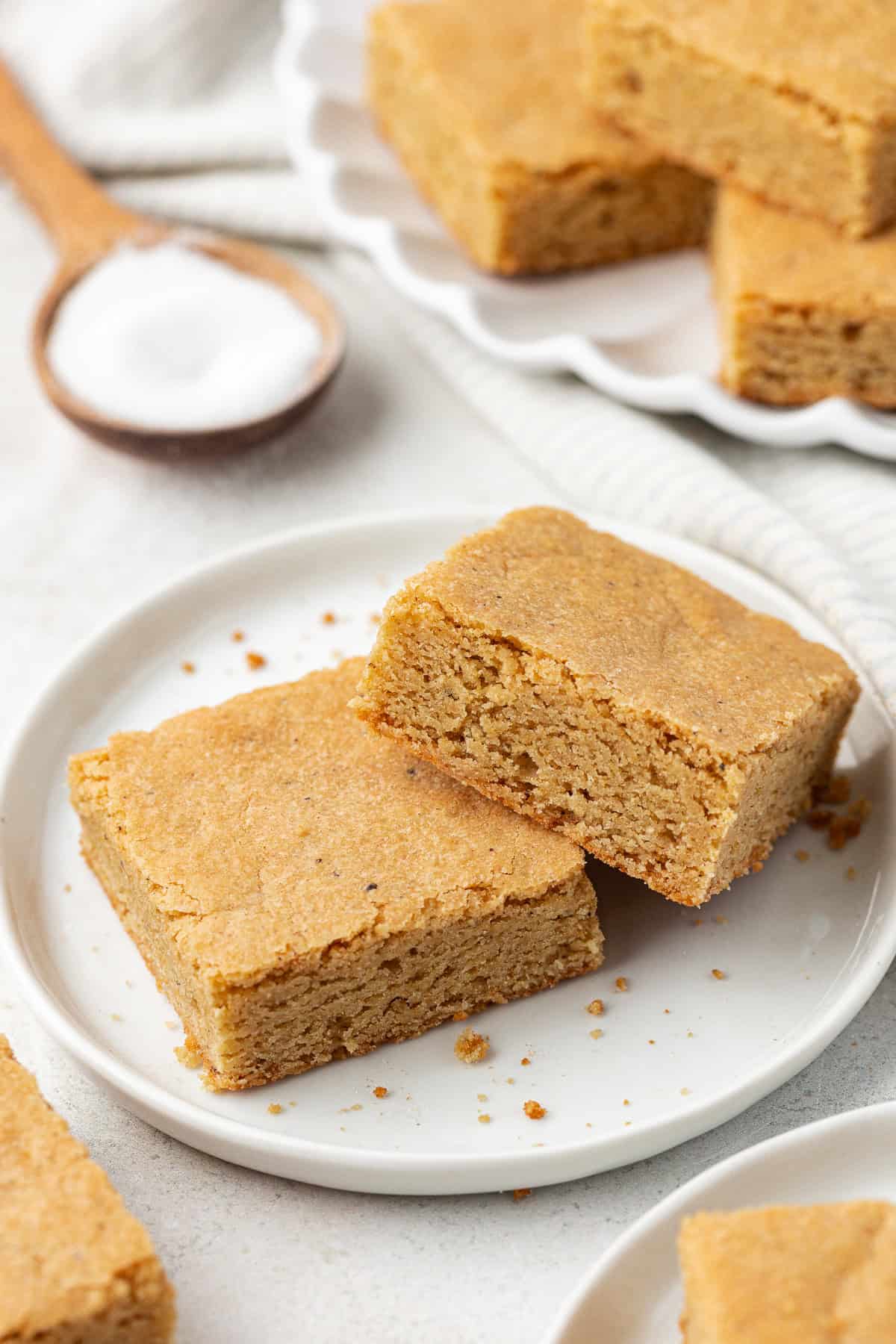 A small white plate with two blondie squares on it surrounded by more blondies on plates and a wood spoon full of sugar