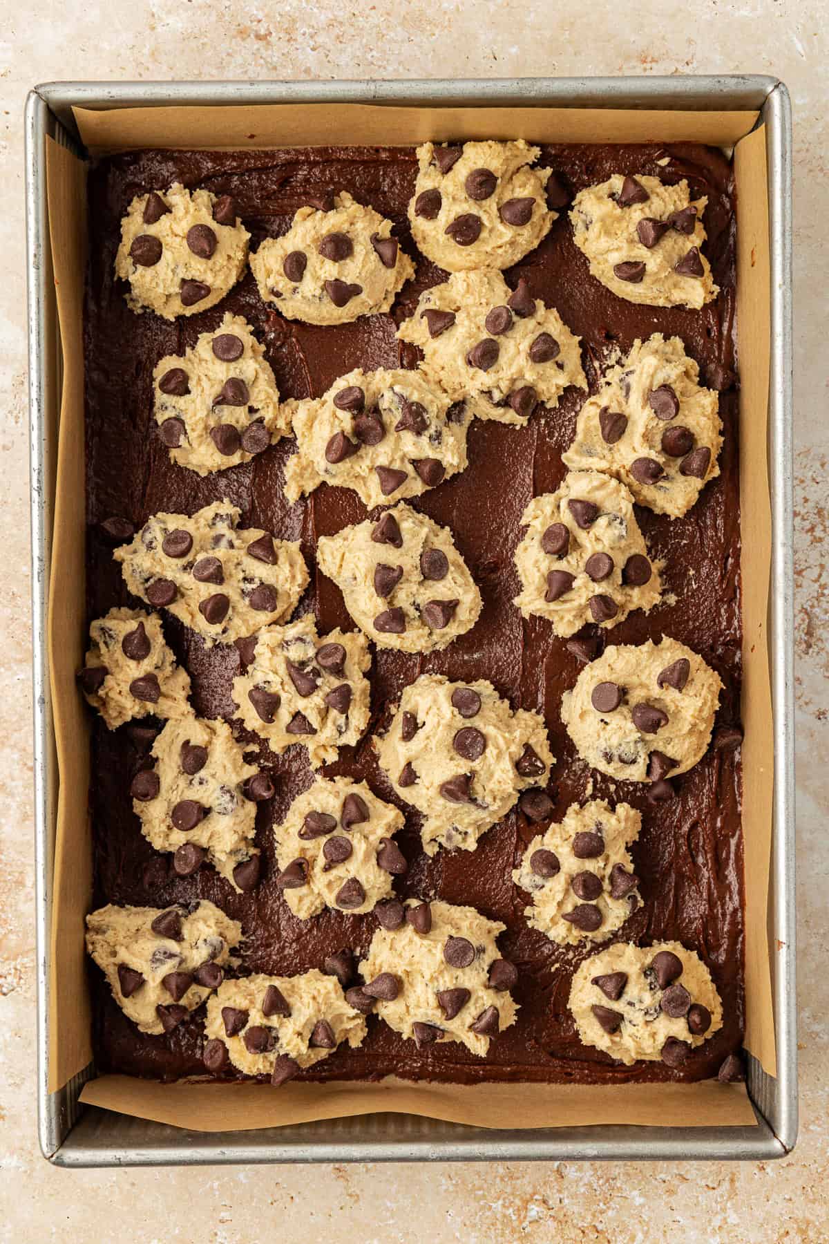 overhead view of a metal baking pan lined with parchment paper with brown batter in the bottom and dollops of chocolate chip cookie dough on top