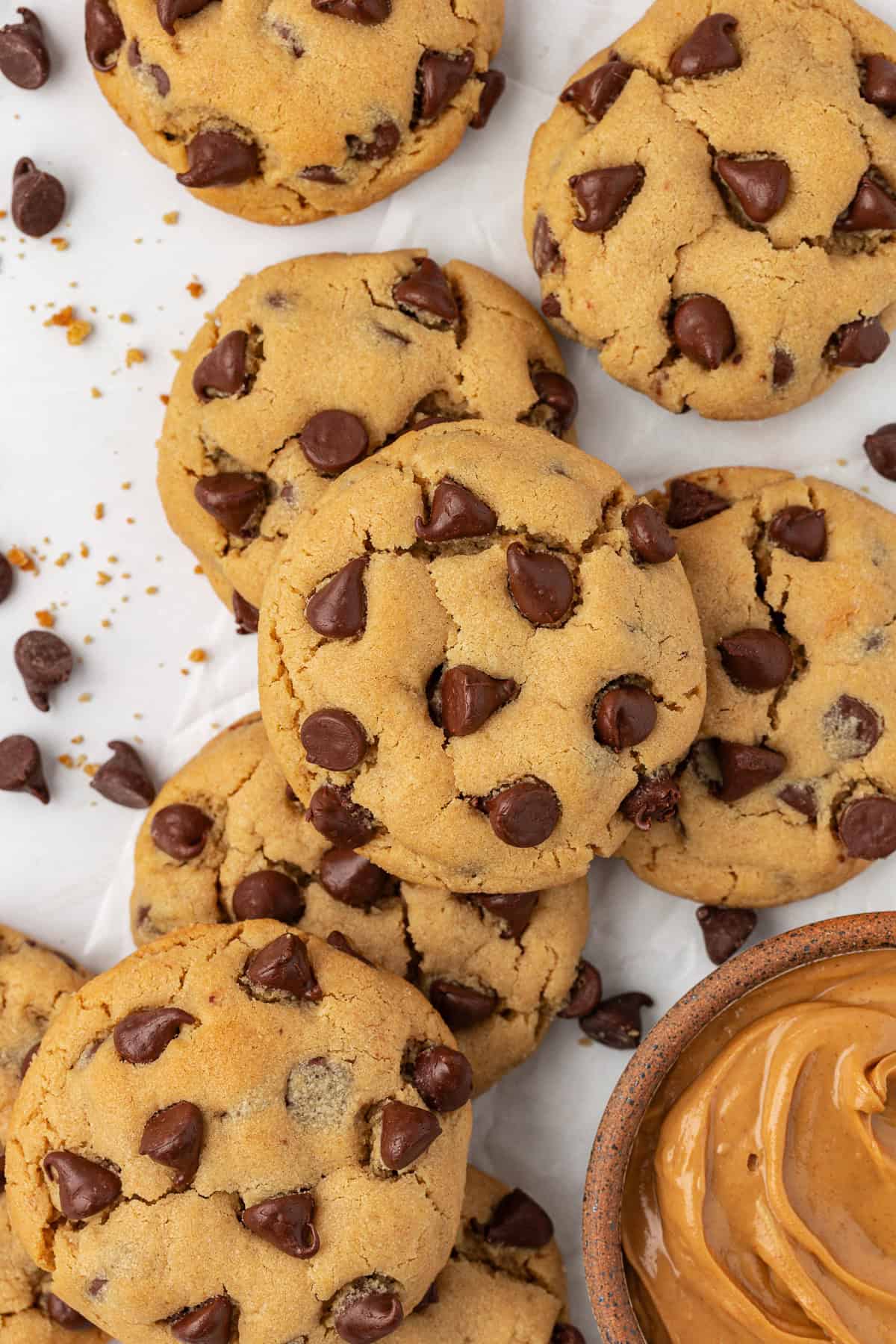 over head view of scattered peanut butter chocolate chip cookies beside a bowl of peanut butter, with chocolate chips and cookie crumbs scattered around