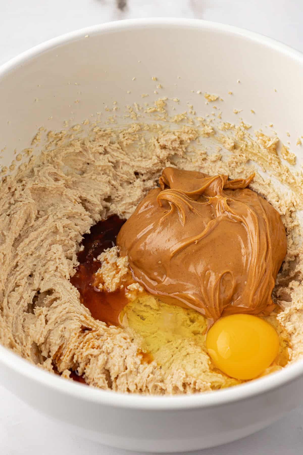 ingredients for peanut butter chocolate chip cookie dough in a white bowl, not combined yet so you see the whole egg, scoop of peanut butter and more liquid ingredients on top