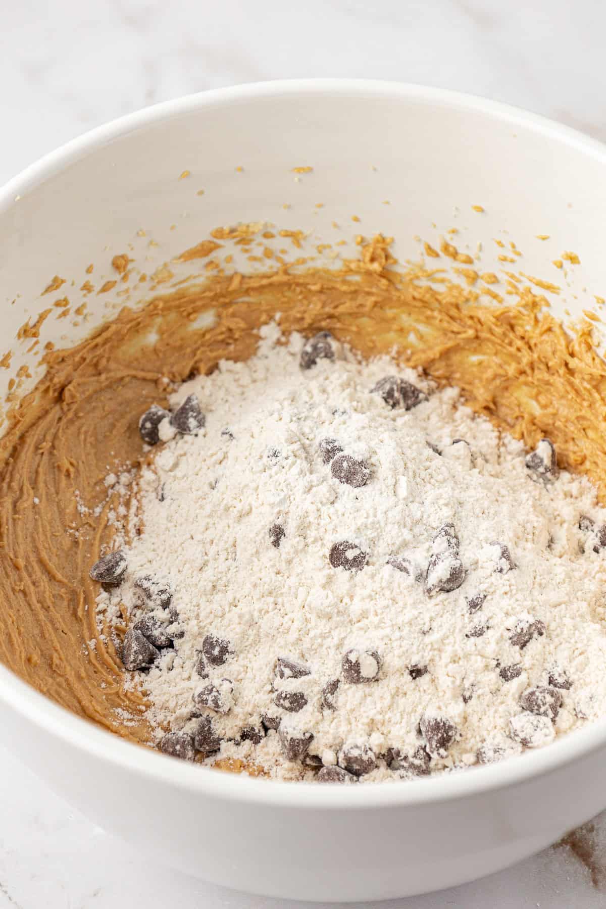 peanut butter chocolate chip cookie dough ingredients with wet ingredients on the bottom and dry ingredients on top, not yet mixed in, in a large white bowl