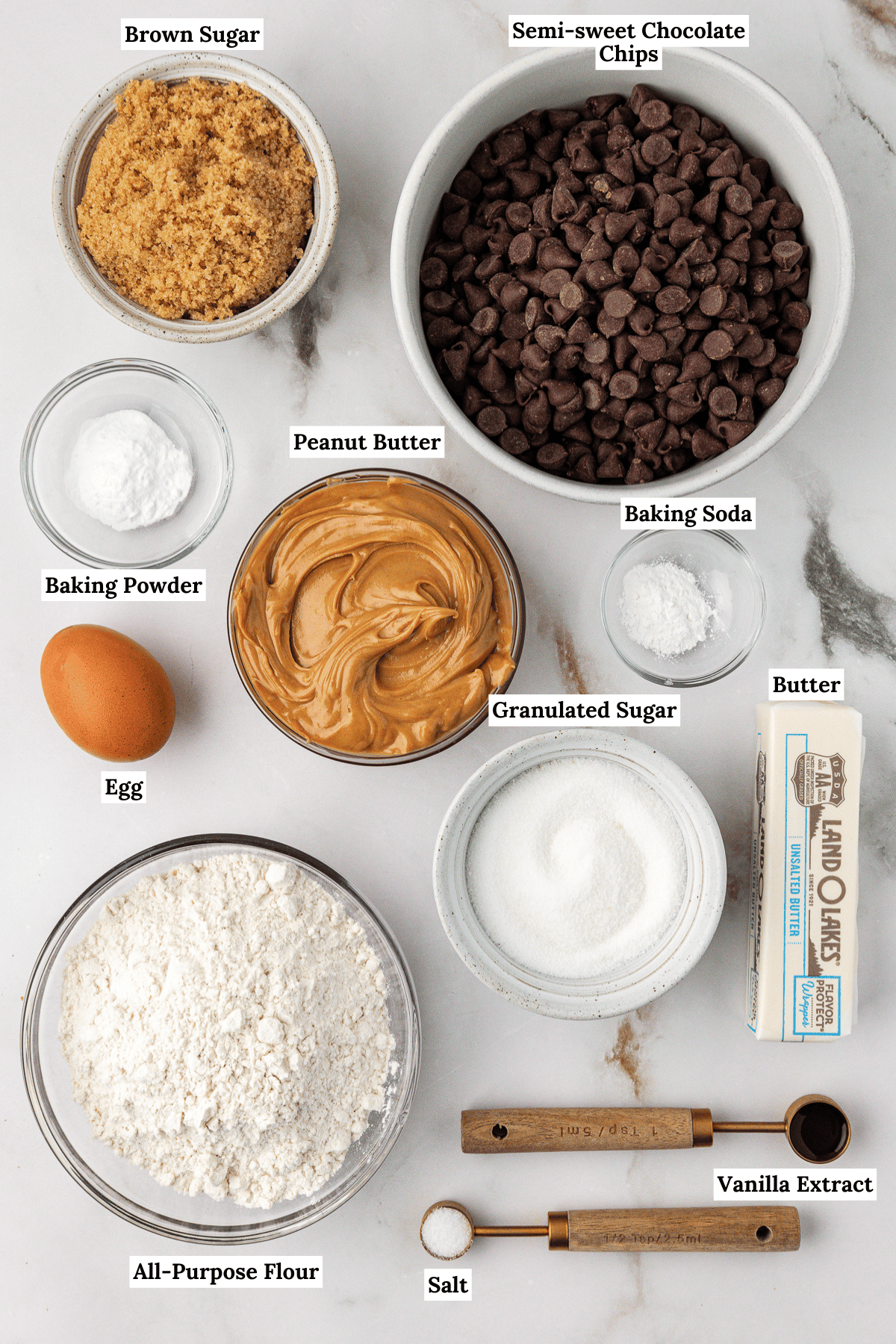 over head view of the ingredients for peanut butter chocolate chip cookies including brown sugar, semi-sweet chocolate chips, baking powder, peanut butter, baking soda, egg, granulated sugar, butter, all-purpose flour, salt and vanilla extract