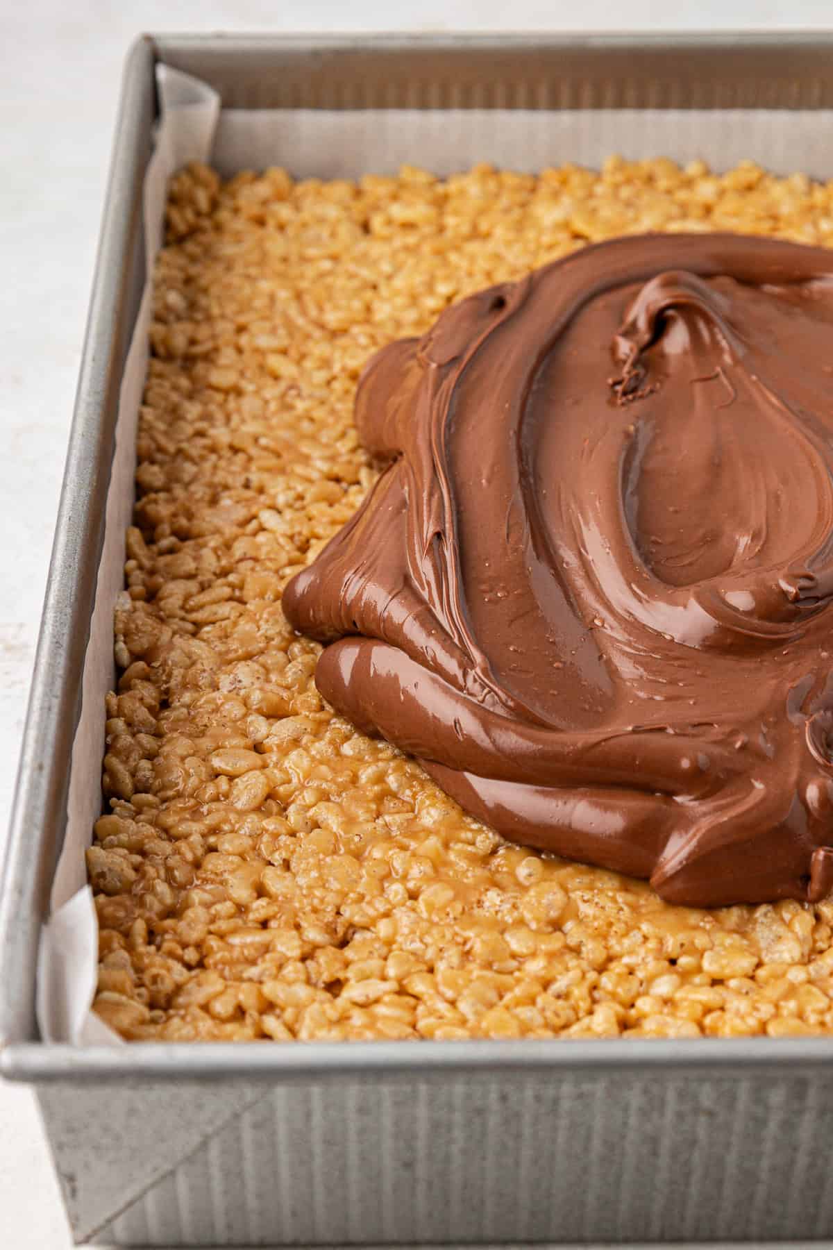 a baking dish lined with parchment paper and packed with a rice krispie mixture, topped with a large dollop of a melted chocolate mixture